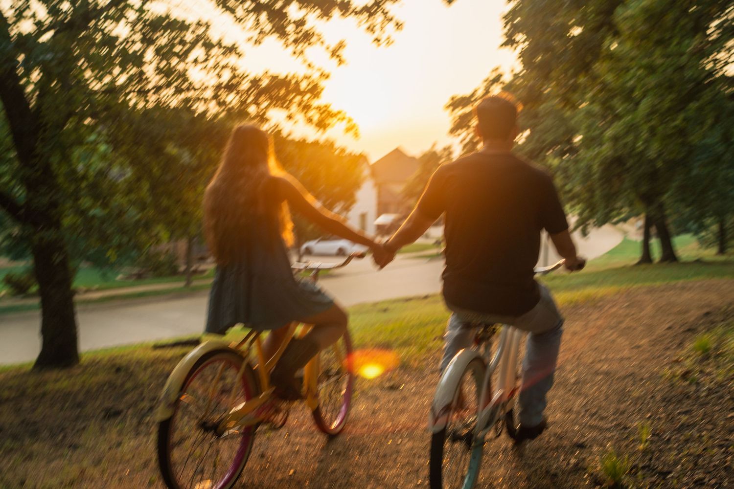 Two cyclists riding bikes together along a path at sunset while holding hands in a romantic summer scene.