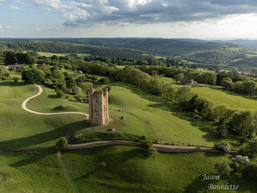 A scenic view of Broadway Tower, a historic folly atop the Cotswolds hills, surrounded by rolling countryside under a partly cloudy sky.