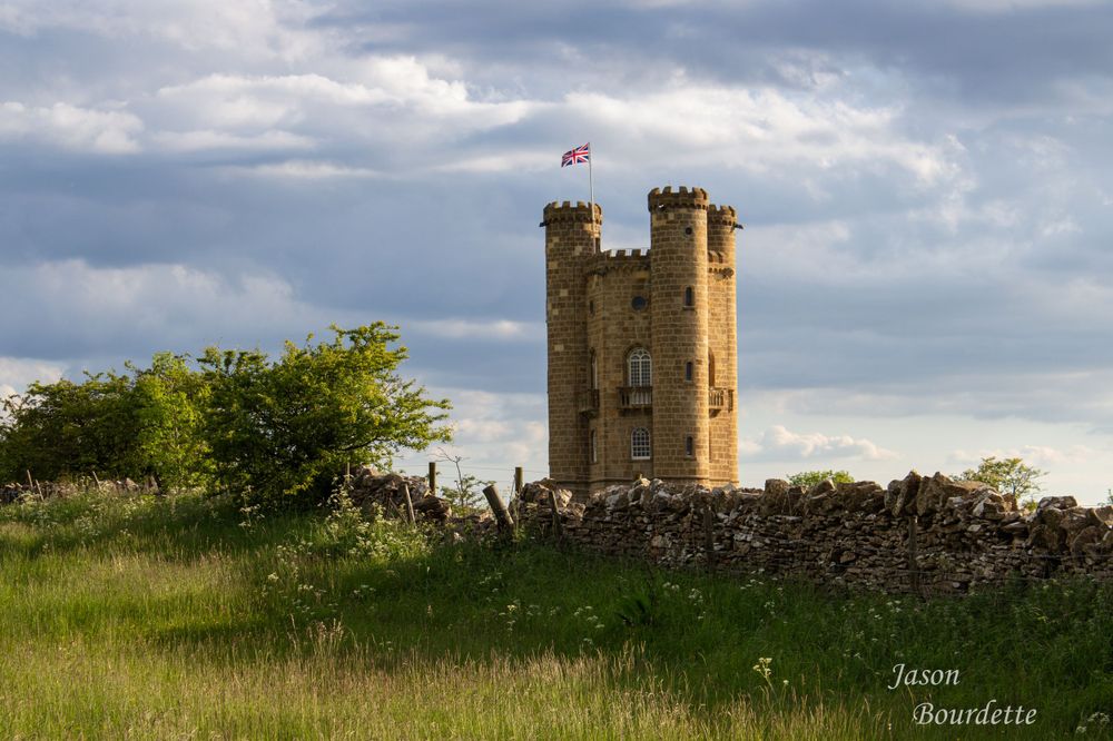 A scenic view of Broadway Tower, a historic folly atop the Cotswolds hills, surrounded by rolling countryside under a partly cloudy sky.