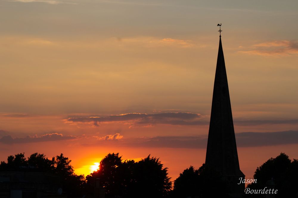 A majestic church steeple rises above the tree line, silhouetted against a stunning sunset. The warm hues of a orange, pink, and purple sky