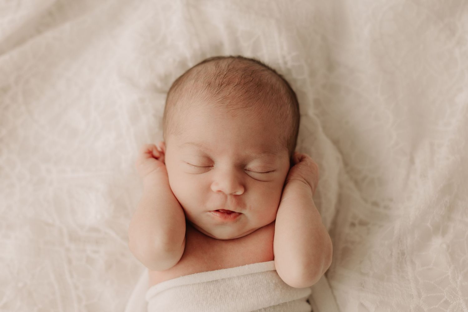 Peaceful newborn sleeping on soft white fabric with hands near face.