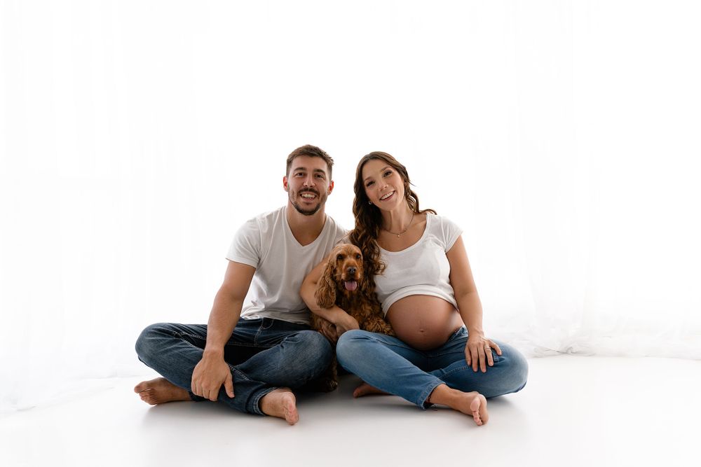 Couple sits cross-legged with their dog during maternity photoshoot against white backdrop.