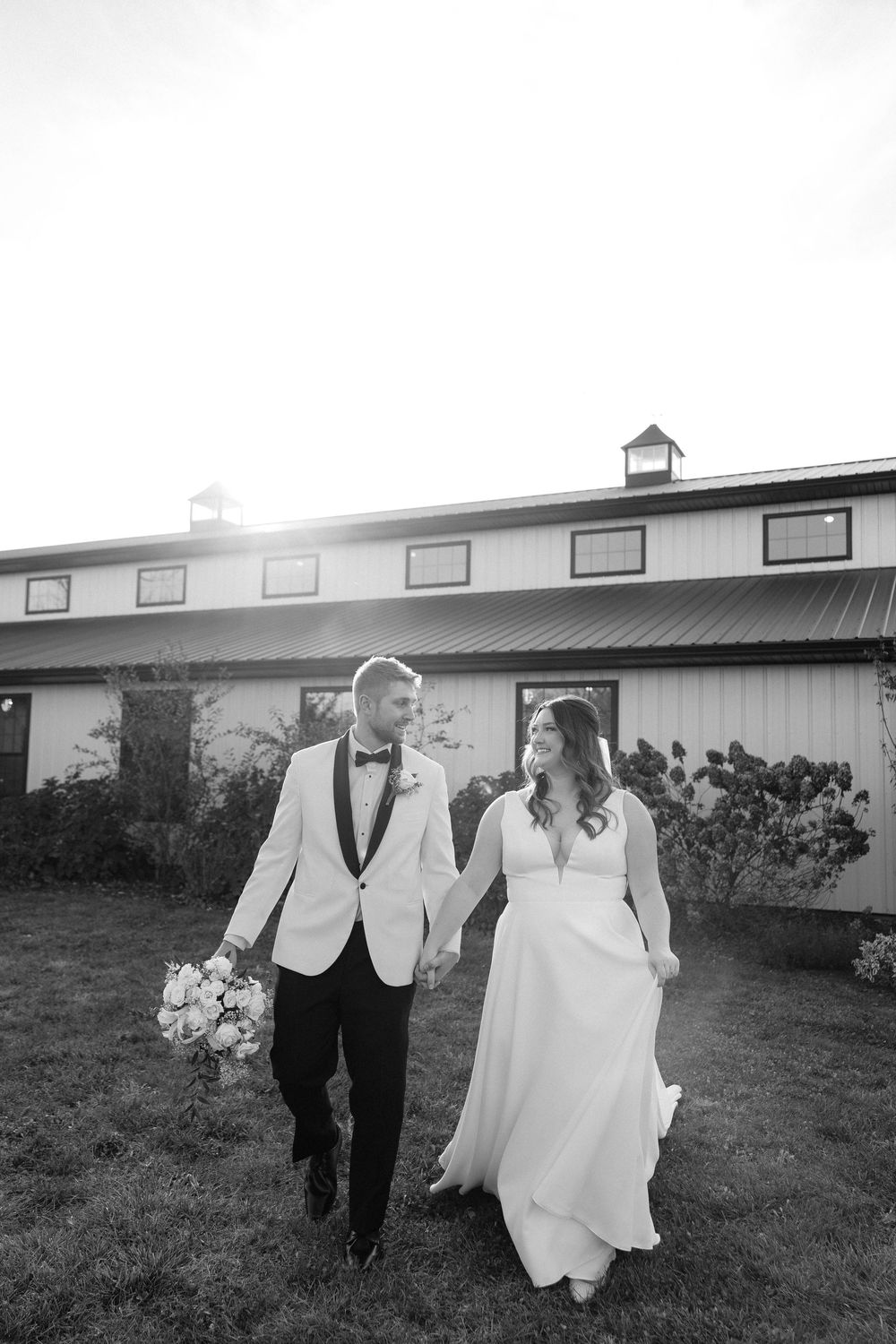 Newlywed couple runs playfully through a grassy field outside a rustic barn on their wedding day.