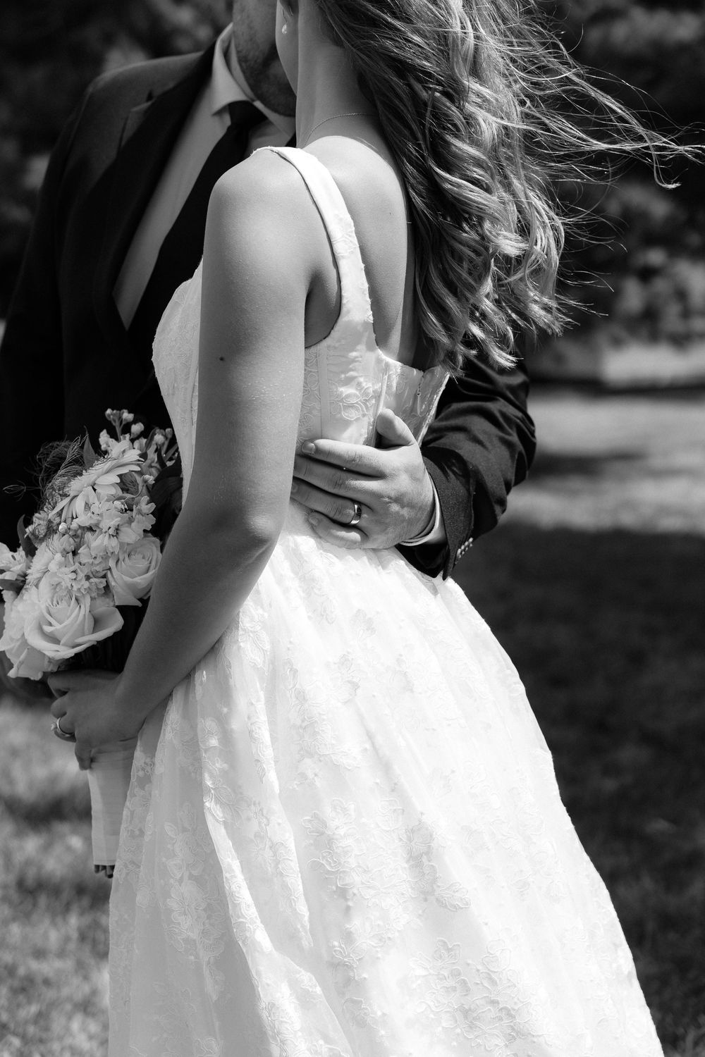 Black and white wedding photo of bride in lace gown holding bouquet, embraced by groom in suit outdoors.