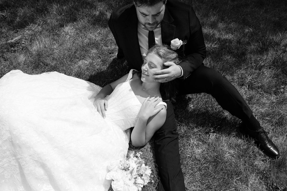 Black and white wedding photo of bride and groom sitting on grass, sharing a tender romantic moment.