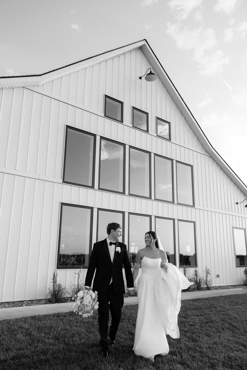 Black and white photo of a bride and groom walking hand in hand in front of a modern white barn wedding venue.