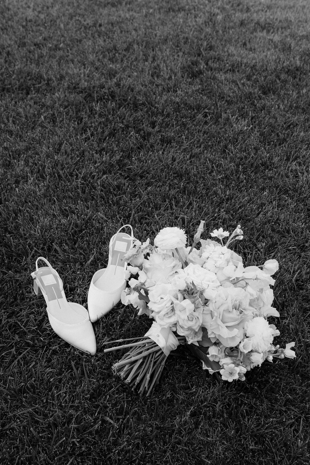 Black and white photo of bridal bouquet with white flowers and elegant heels on green grass.