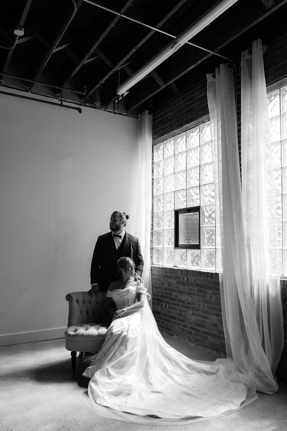 Black and white wedding photo of bride in gown seated in chair with groom standing beside large window in industrial venue.