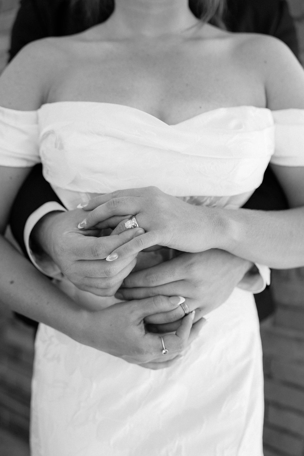 Black and white photo of bride and groom embracing, showing wedding rings on their intertwined hands.