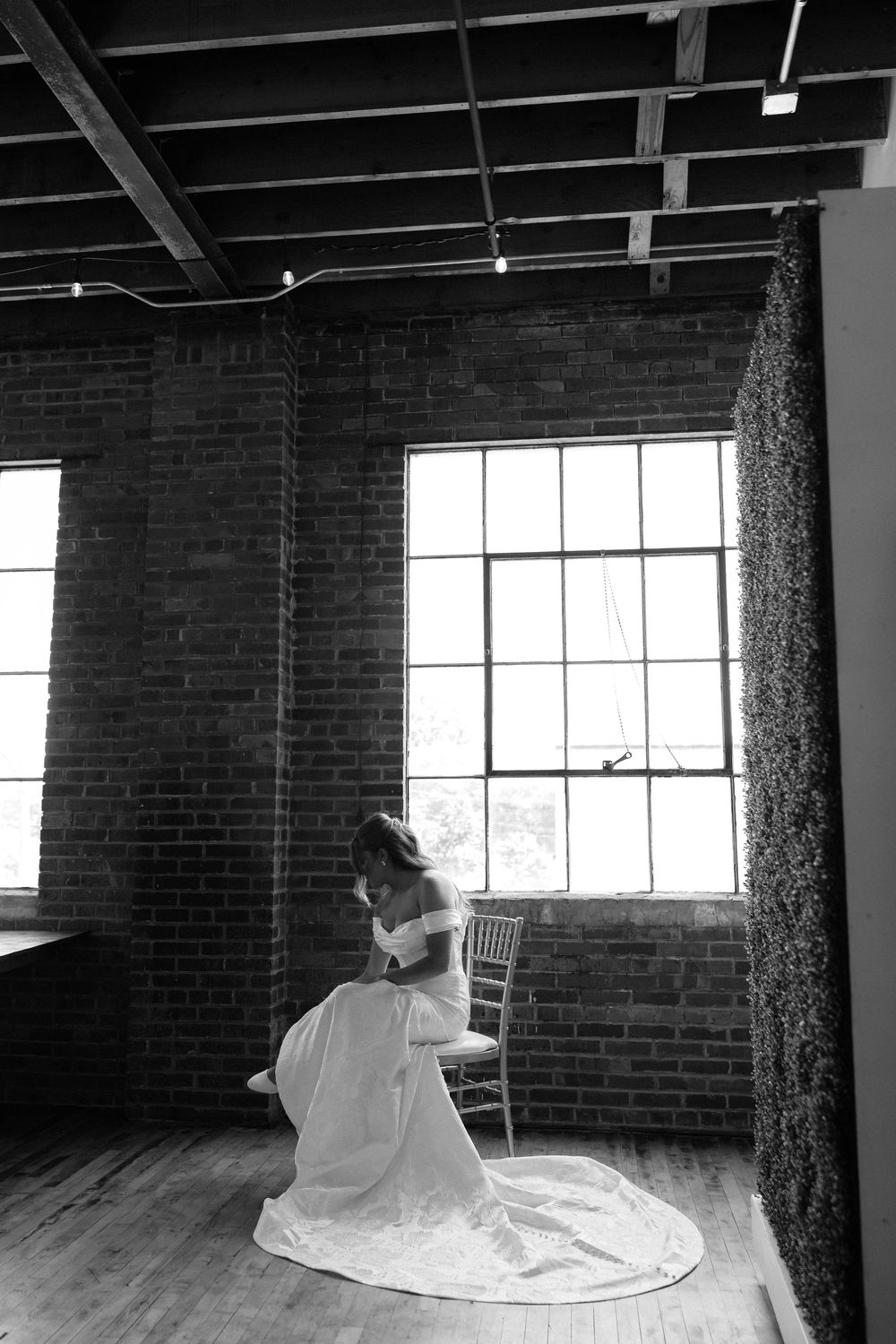 Black and white photo of a bride in a flowing gown seated in a chair inside a rustic brick-walled venue.