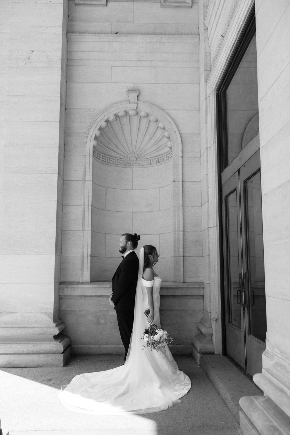 Black and white photo of bride and groom standing before ornate stone arch at elegant wedding venue.