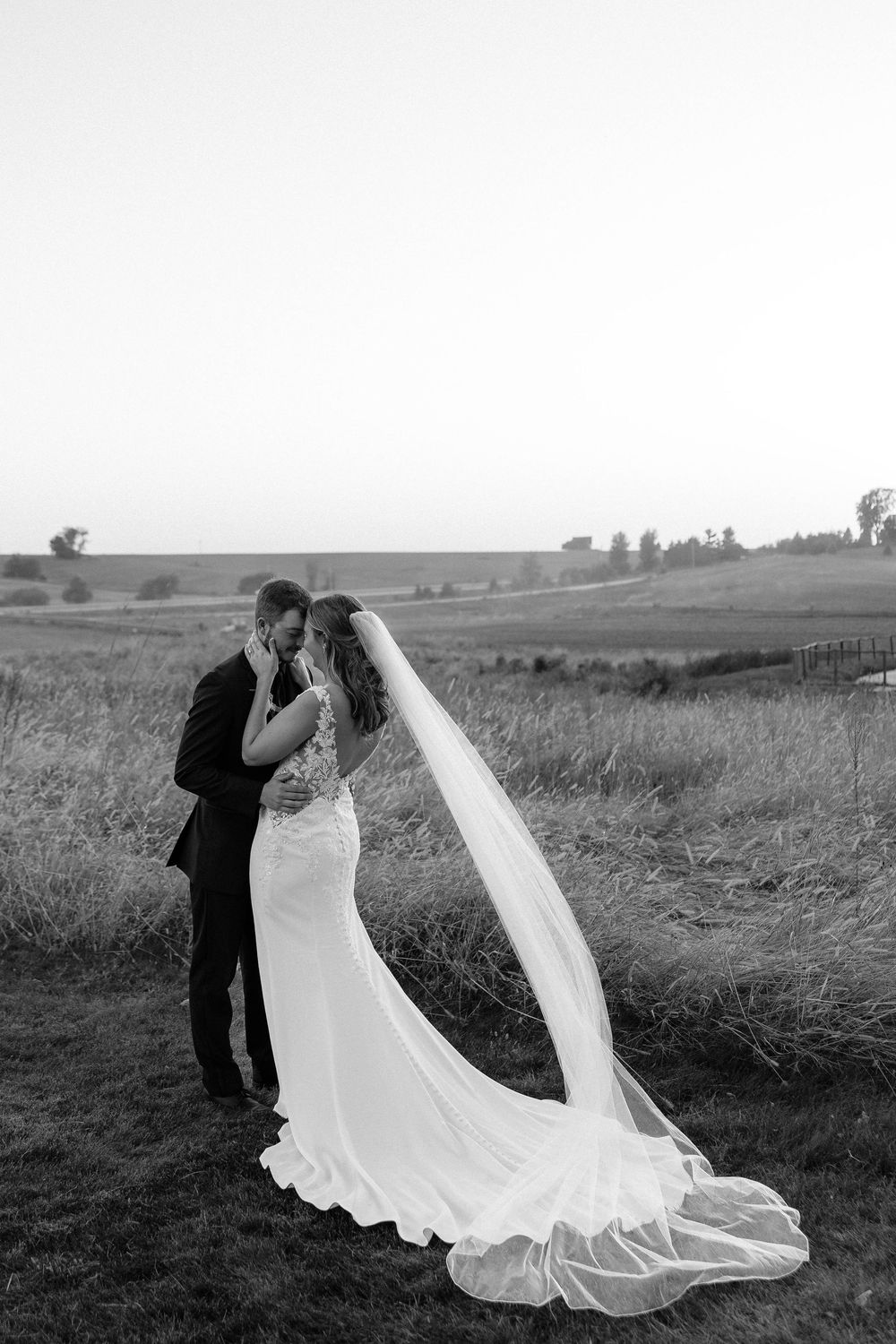 A romantic black and white wedding photo showing a couple embracing on a hilltop with a long flowing veil in the breeze.