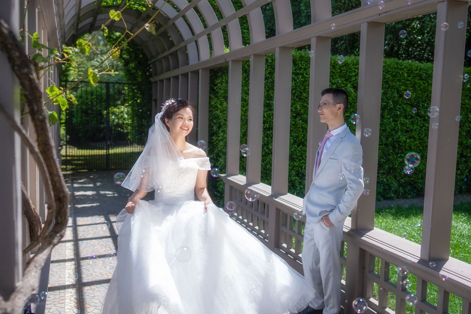 Bride and groom smiling at each other under a garden pergola surrounded by floating bubbles for romantic wedding portraits.