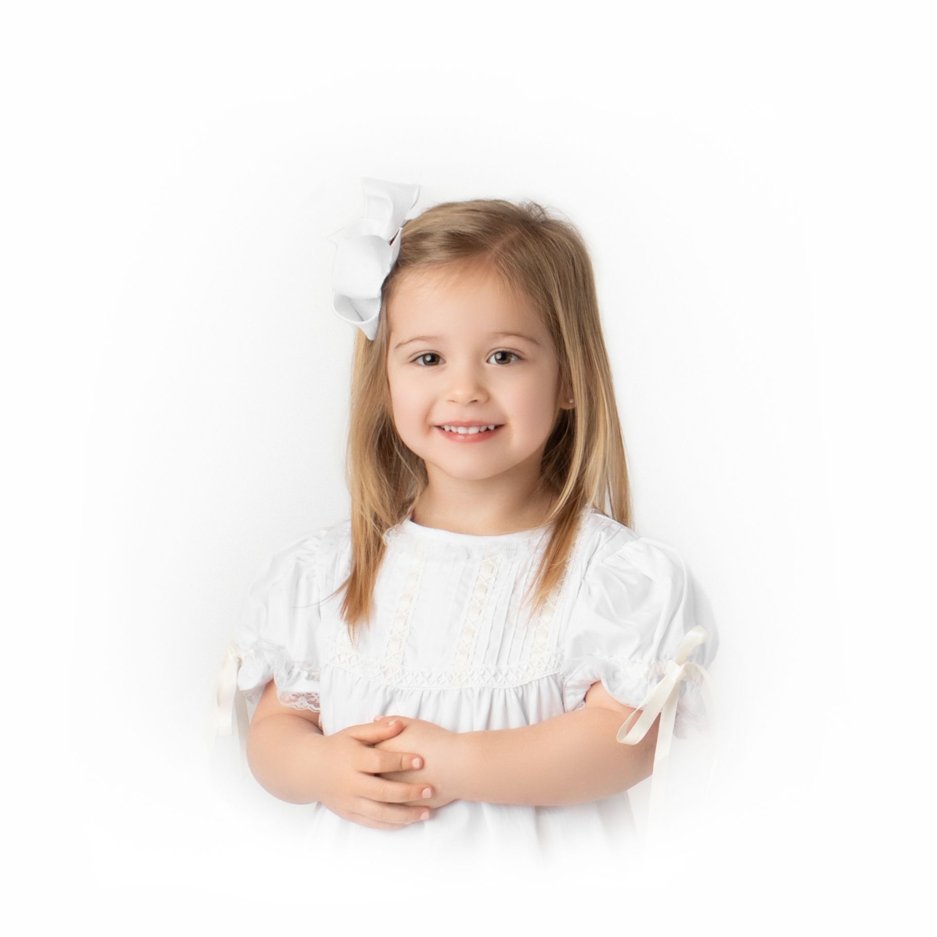 Child in white dress poses sweetly against light background in portrait session.