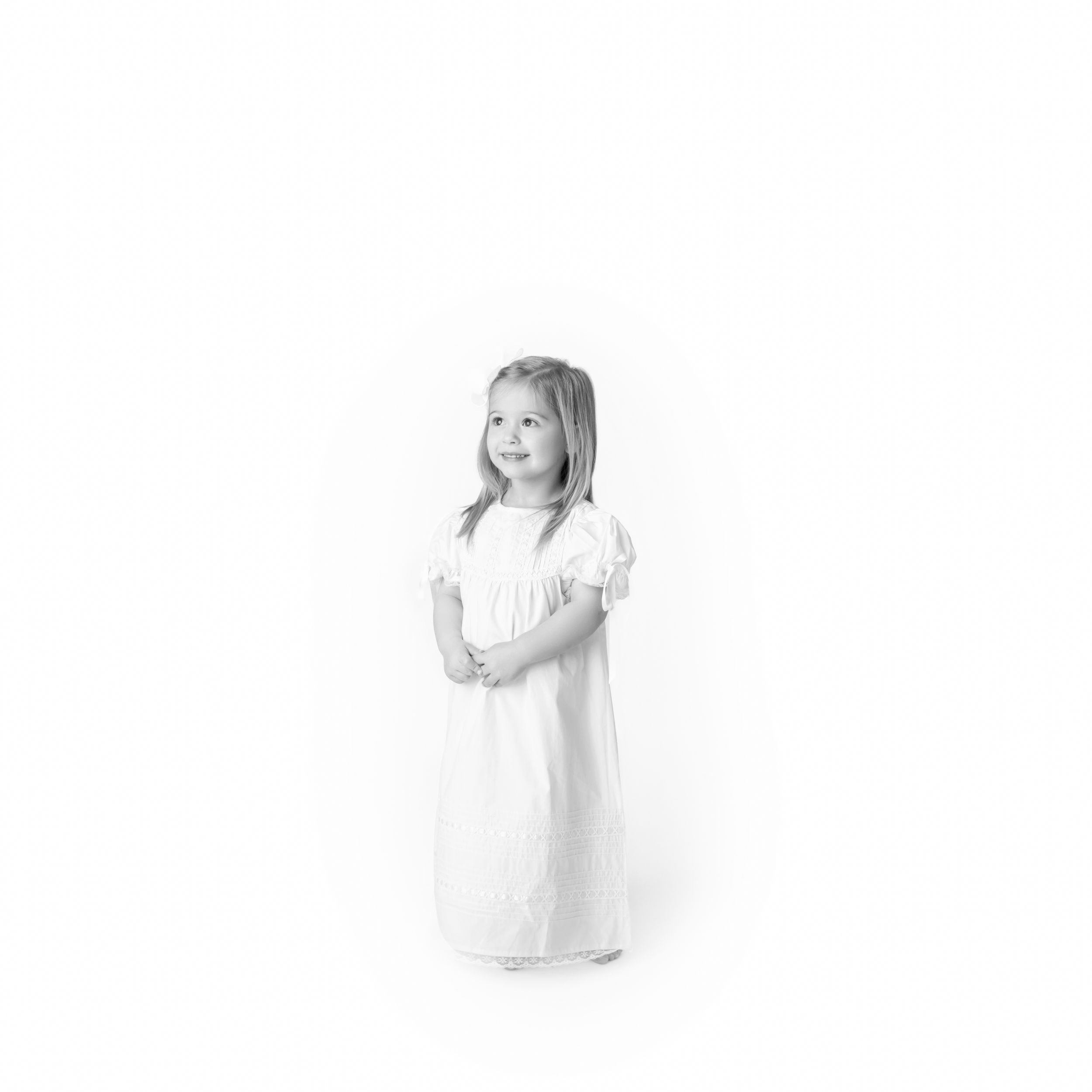 Child wearing white dress stands against soft white background in minimalist portrait.
