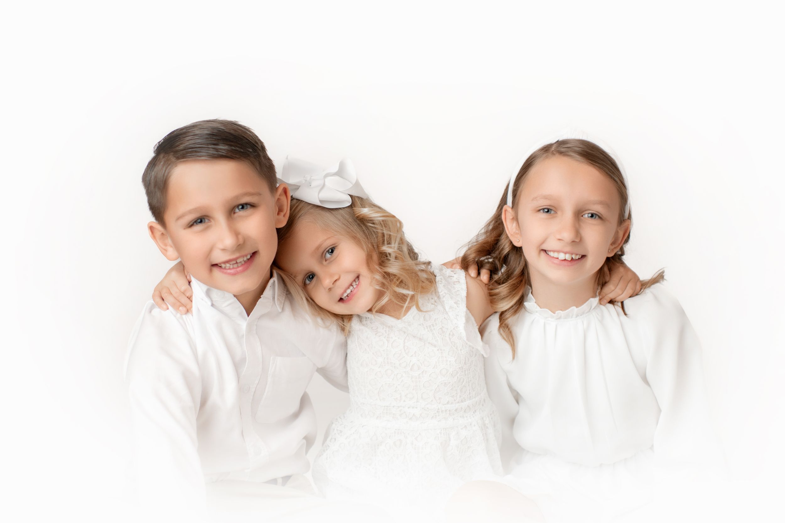 Three children in white clothing pose together against white background.