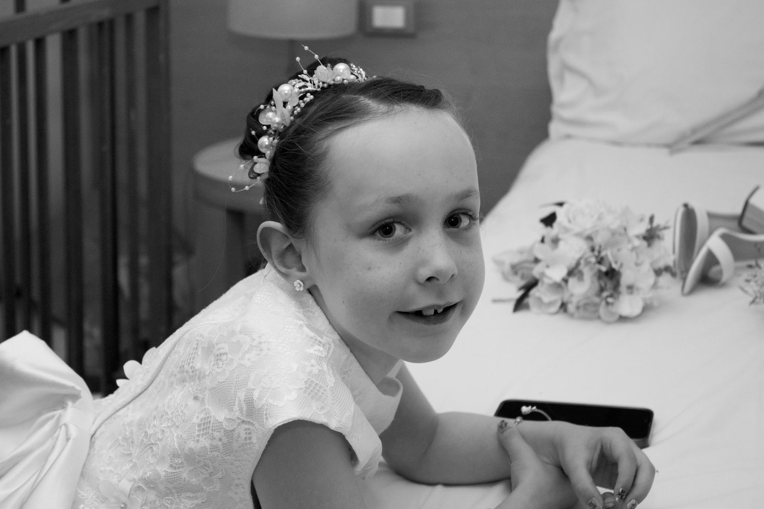 A child in white lace dress and tiara smiles while lying down.