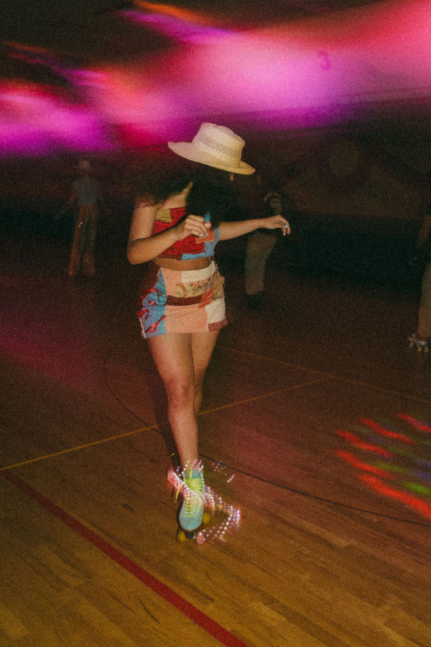 A roller skater in a hat and casual outfit glides on a wooden floor under pink and purple lighting.