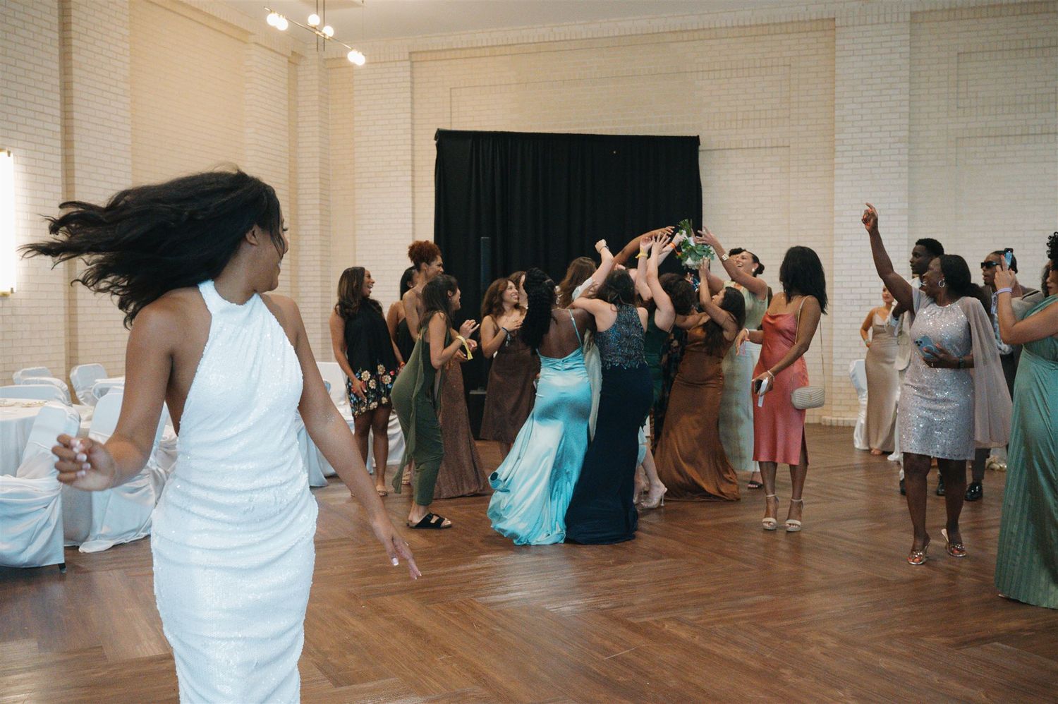 Energetic dancers move on a wooden floor at a celebratory event in a bright room with white walls.