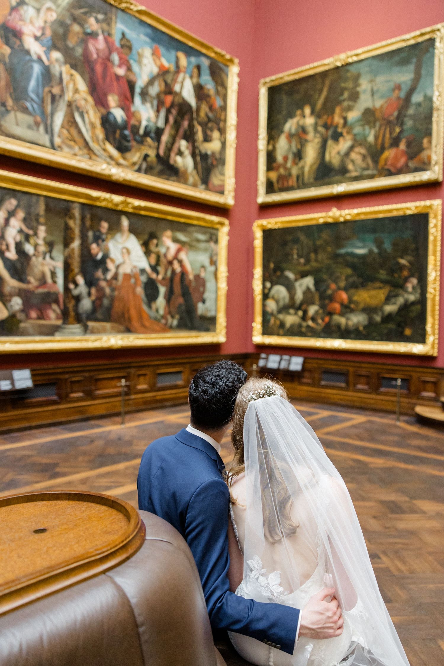 Two people sit on a bench admiring classical paintings in an ornate museum gallery with red walls.