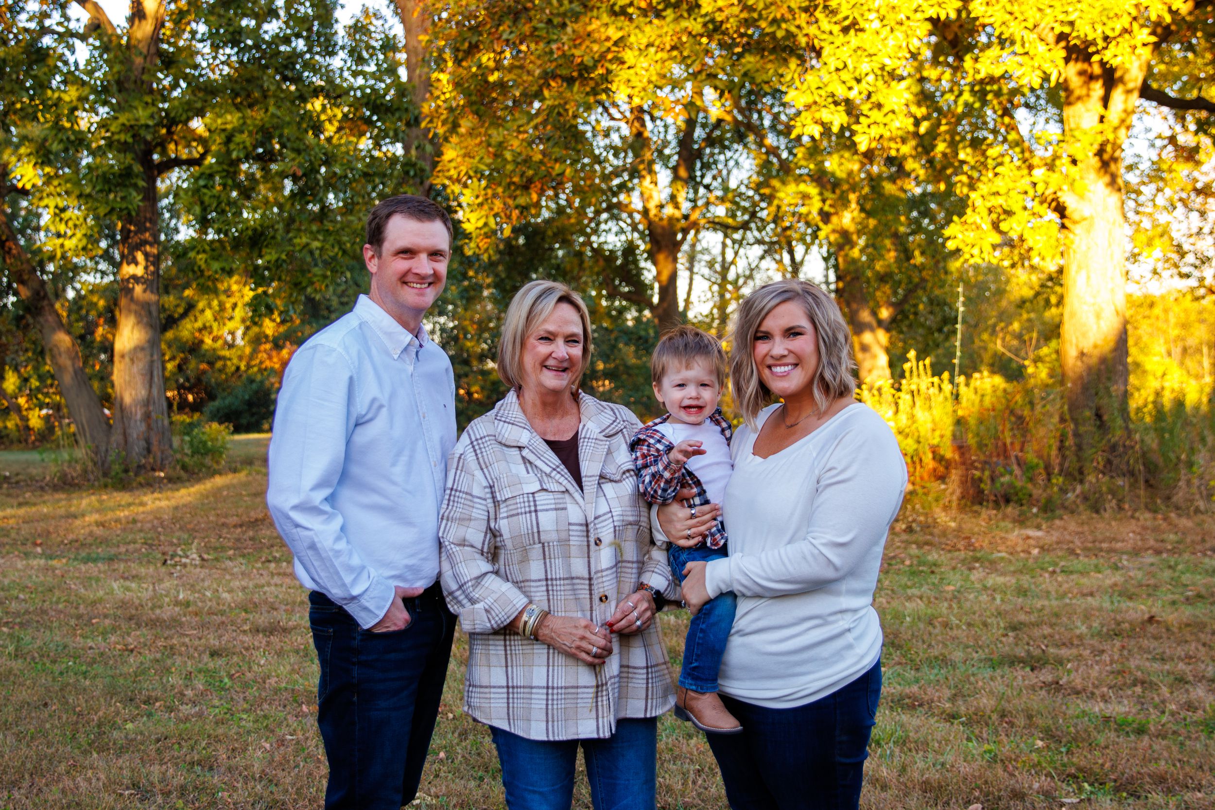 A family poses together outdoors during golden hour with fall foliage in the background.