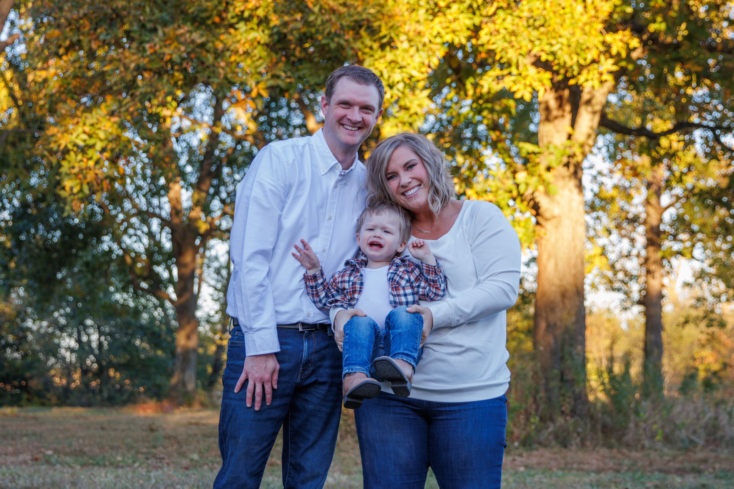 A small family poses together outdoors during sunset with autumn trees in the background.