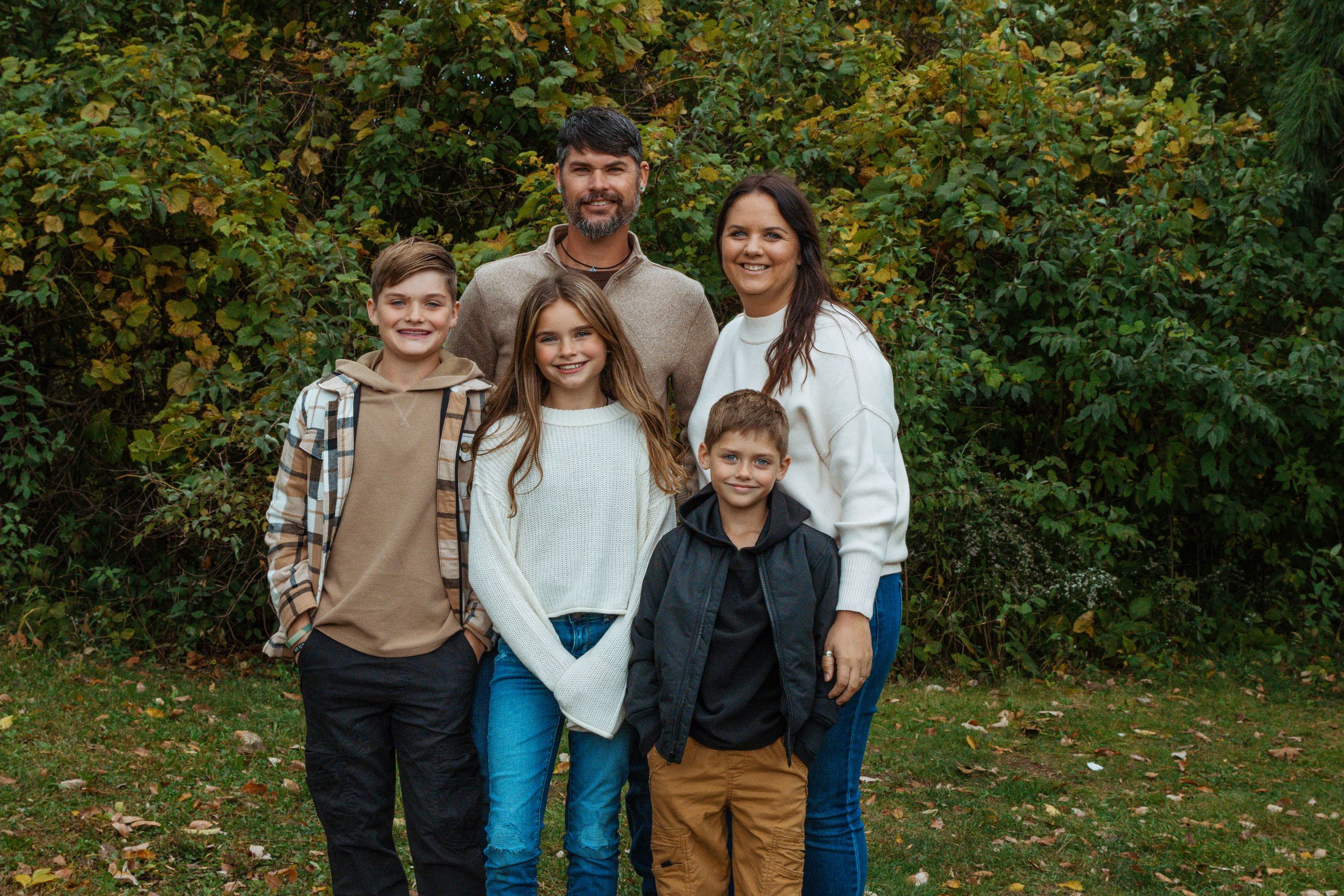 Extended family gathered outdoors for group photo in front of green bushes.