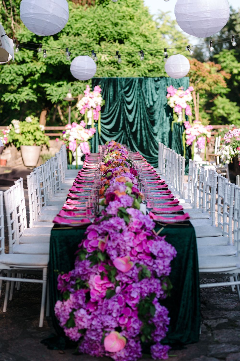 Elegant outdoor wedding reception table setup with purple flowers, white chairs, and hanging paper lanterns in a garden setting.