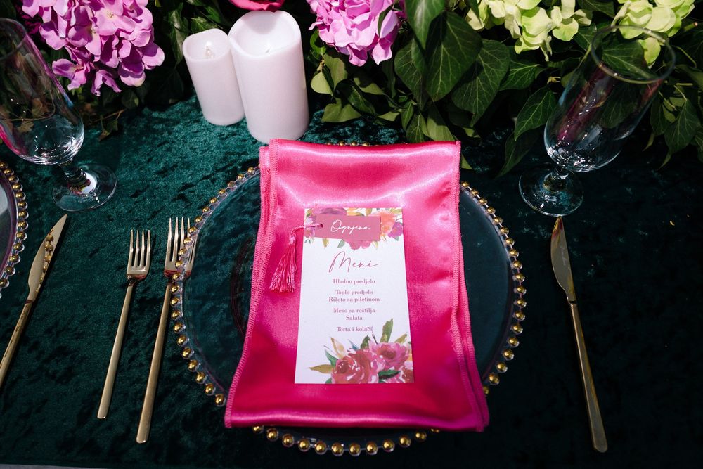 Pink menu card on emerald velvet tablecloth with gold cutlery, glass charger plates, and floral arrangements.