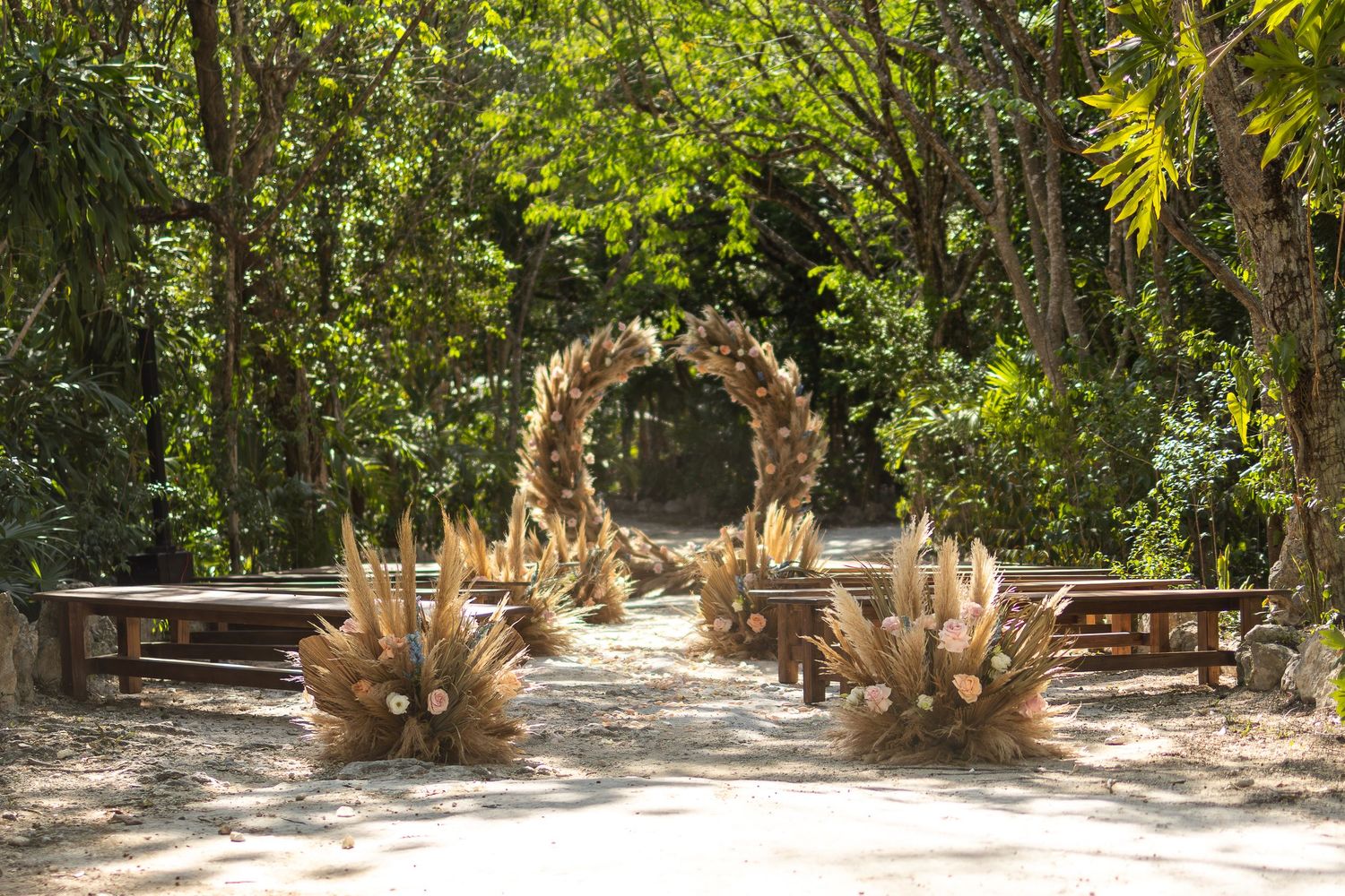 Romantic outdoor wedding ceremony setup with dried palm arch and benches beneath lush greenery.