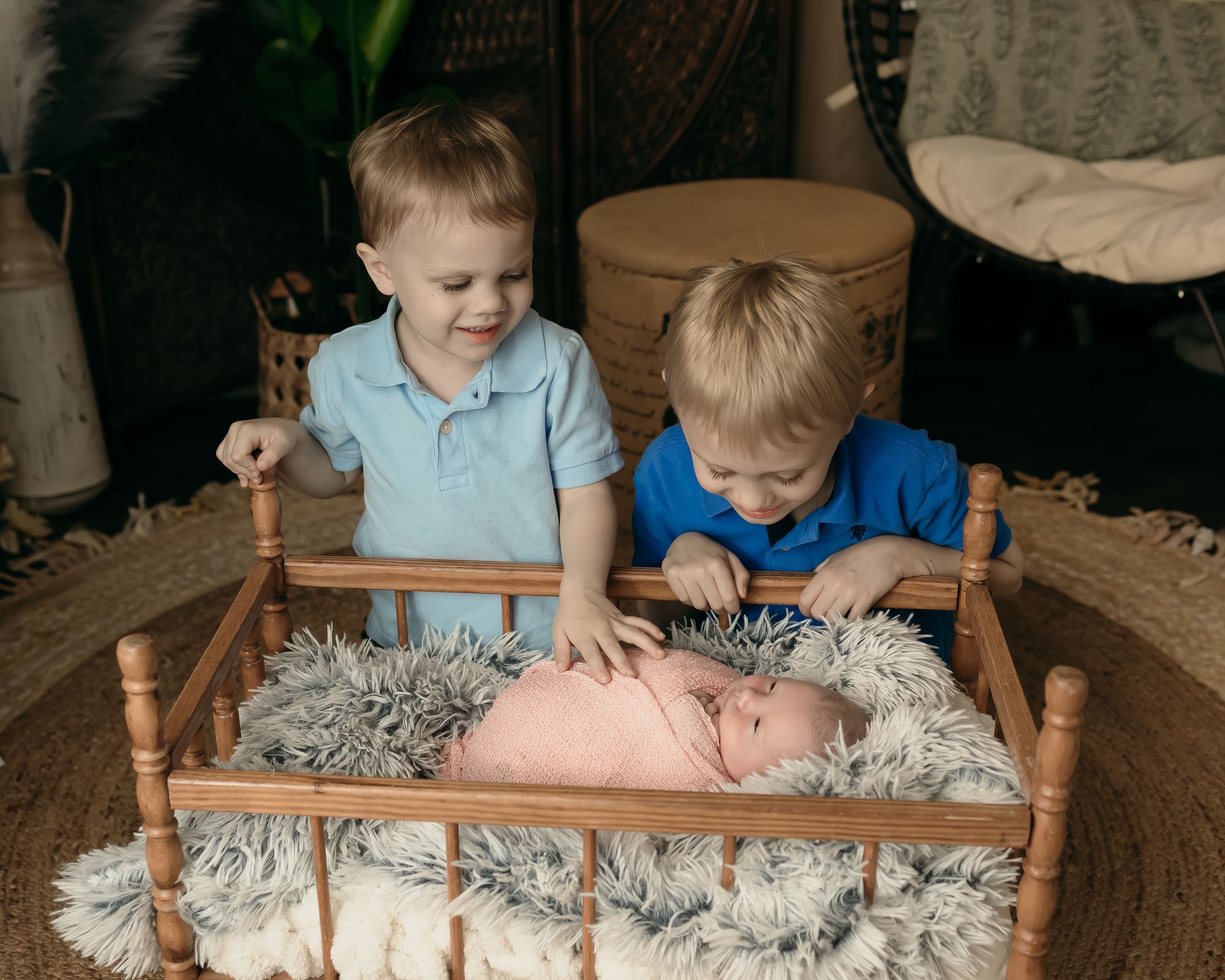 Two young boys admire their newborn sibling resting in a wooden doll bed with fluffy bedding.