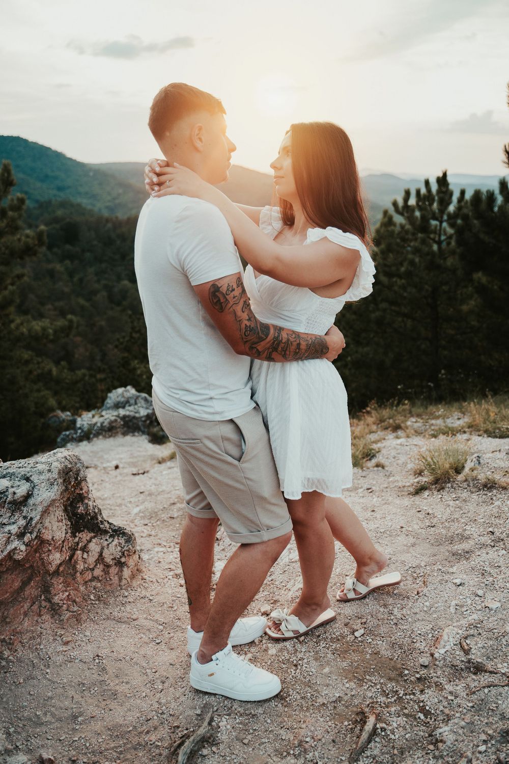 A romantic couple embraces on a mountain cliff during golden hour with scenic mountain views.