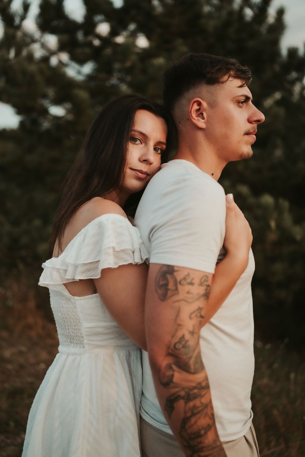 Romantic couple in white outfits pose together at sunset with the woman embracing from behind in a natural setting.