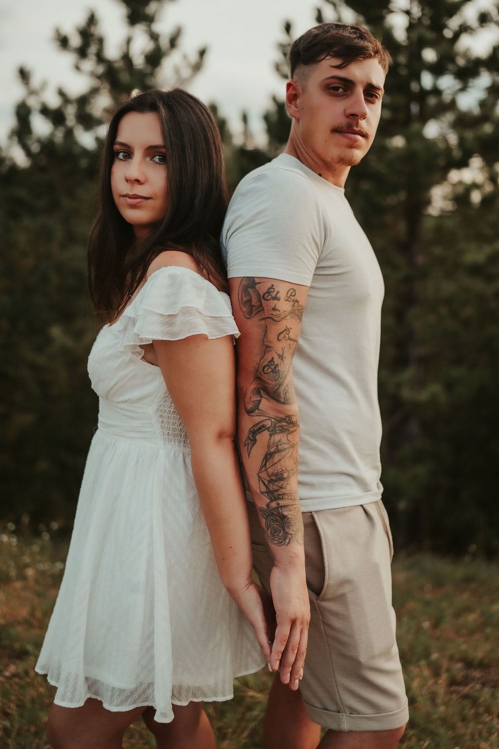 Couple in coordinated white outfits pose back-to-back outdoors with man showing sleeve tattoo.