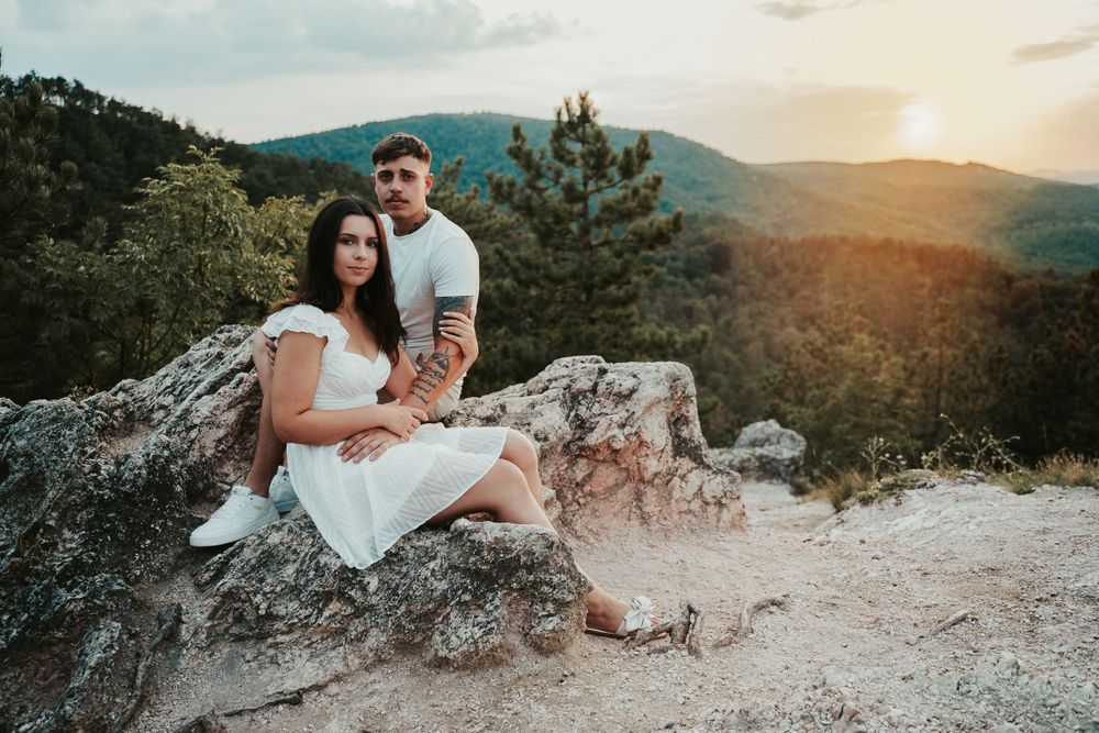 A couple sits together on a rocky overlook at sunset with mountains in the background.