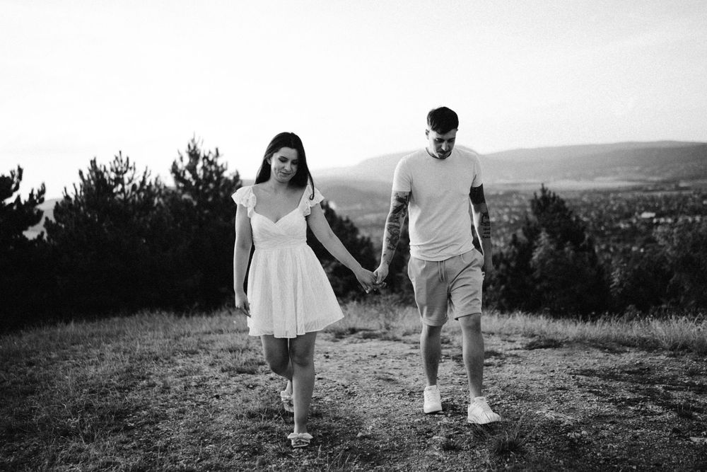 Couple in casual white outfits walking hand in hand on hilltop with scenic mountain view.