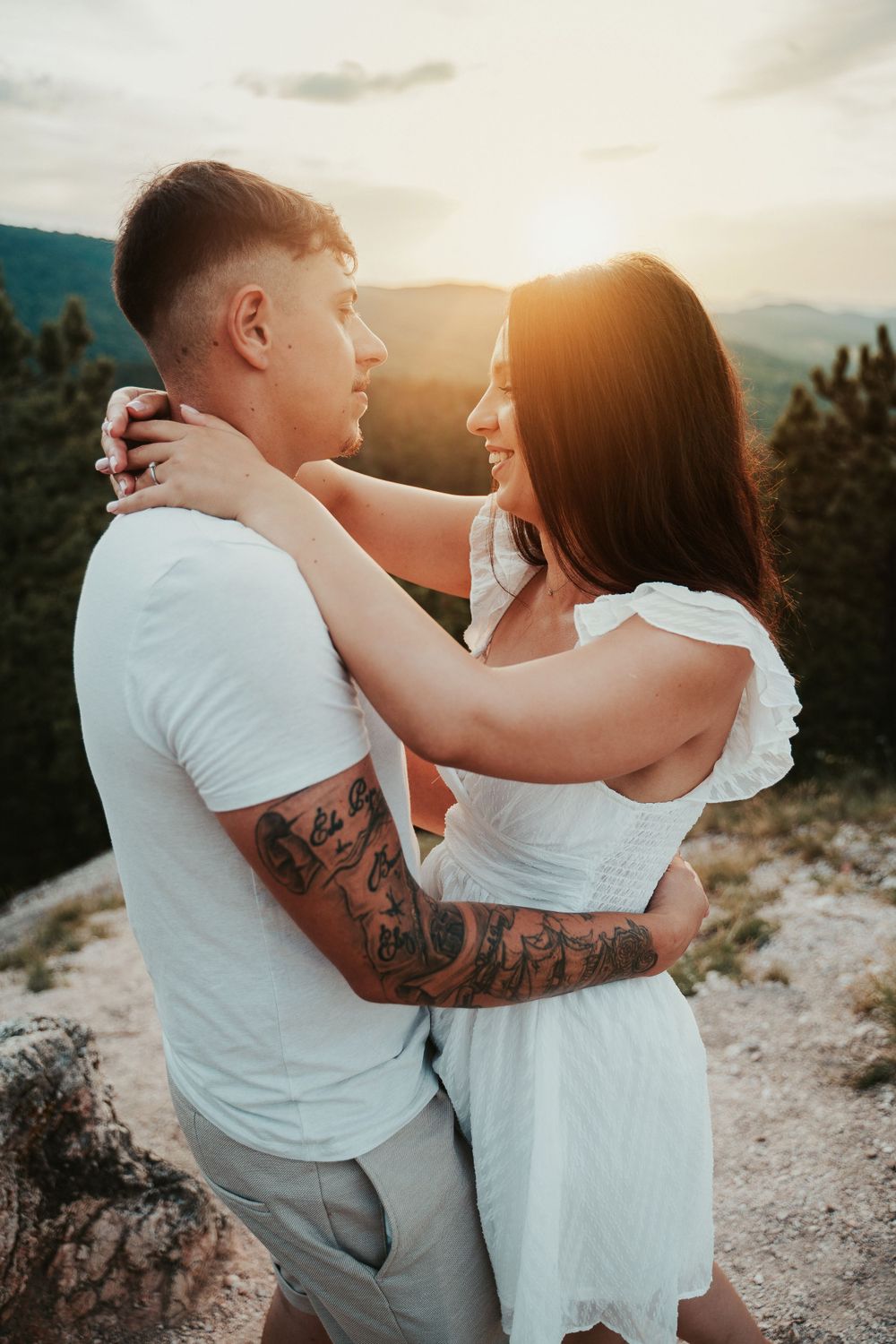 A couple in summer outfits shares a romantic moment against a mountainous sunset backdrop.