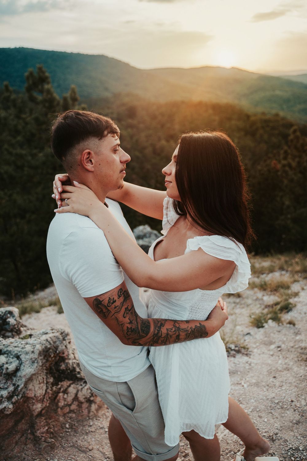 A couple in white and beige outfits embrace on a mountain cliff during sunset.