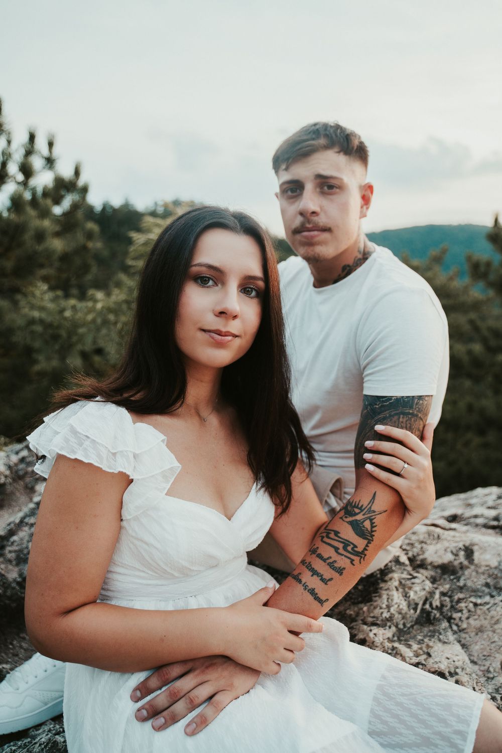 A couple poses for a portrait on a rocky mountain overlook wearing coordinated white outfits.