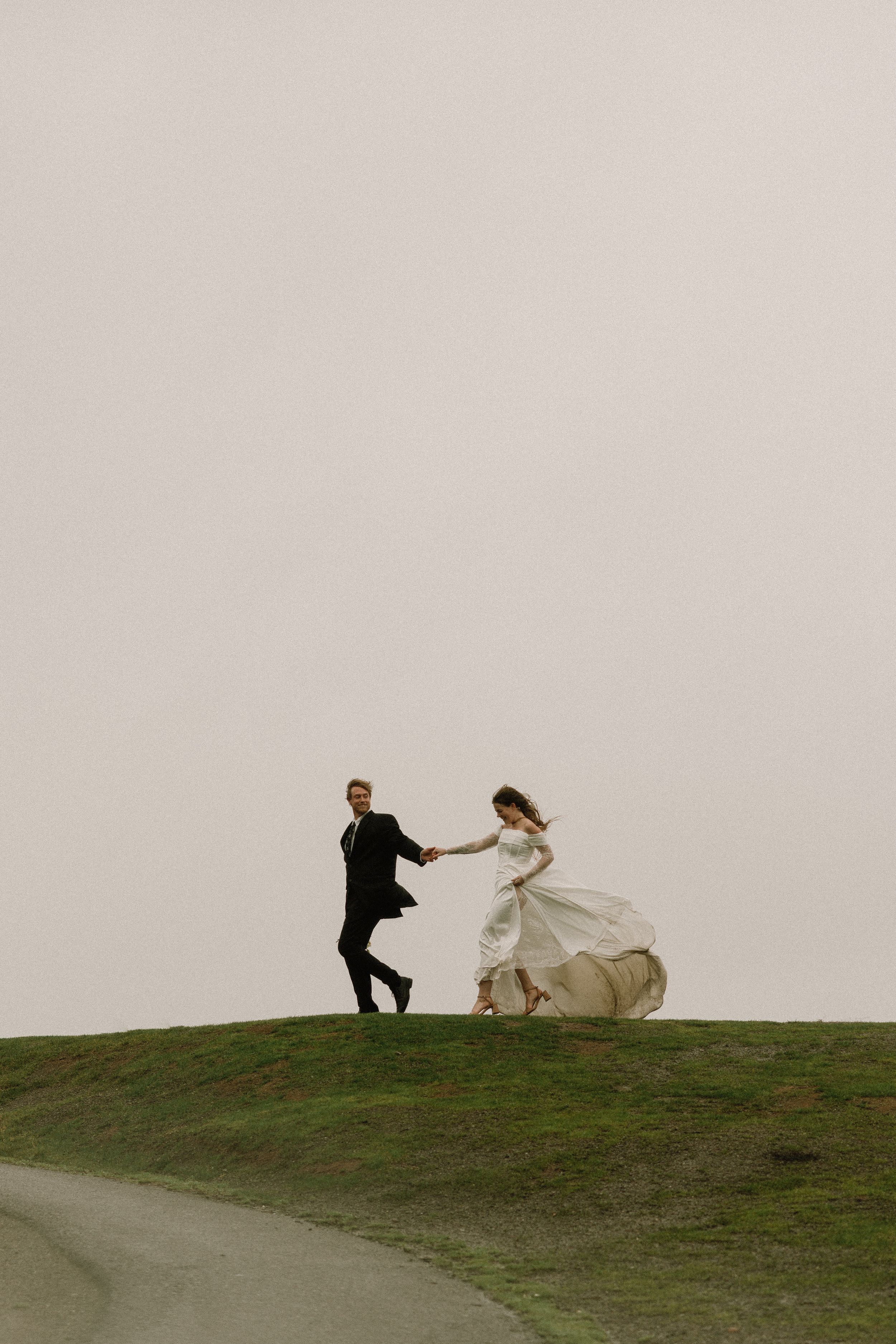 A couple in wedding attire runs playfully across a grassy hilltop silhouetted against an overcast sky.
