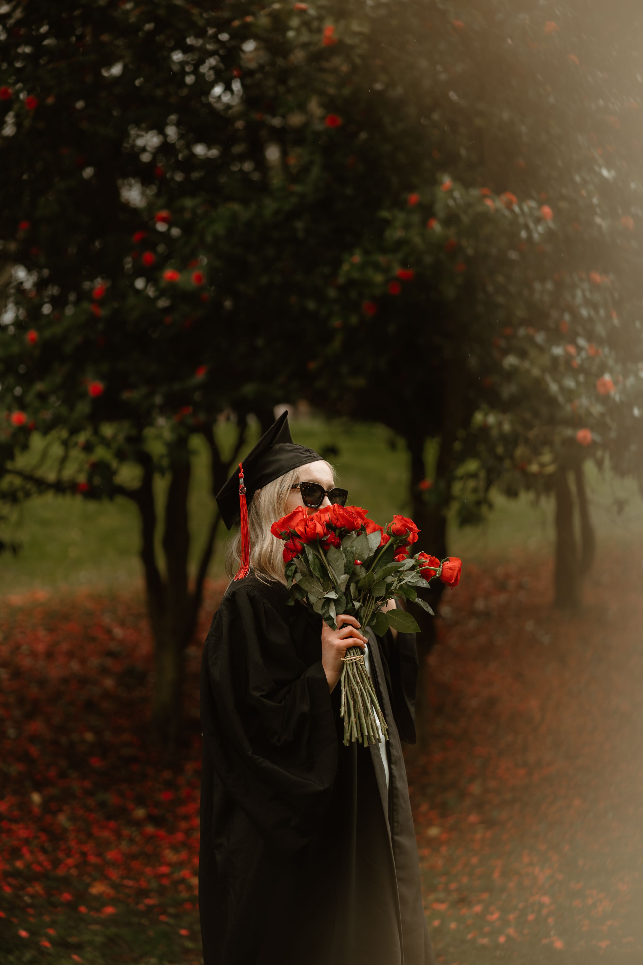 Two figures in black coats embrace in a moody autumn garden with red flowers and fallen leaves creating a romantic scene.