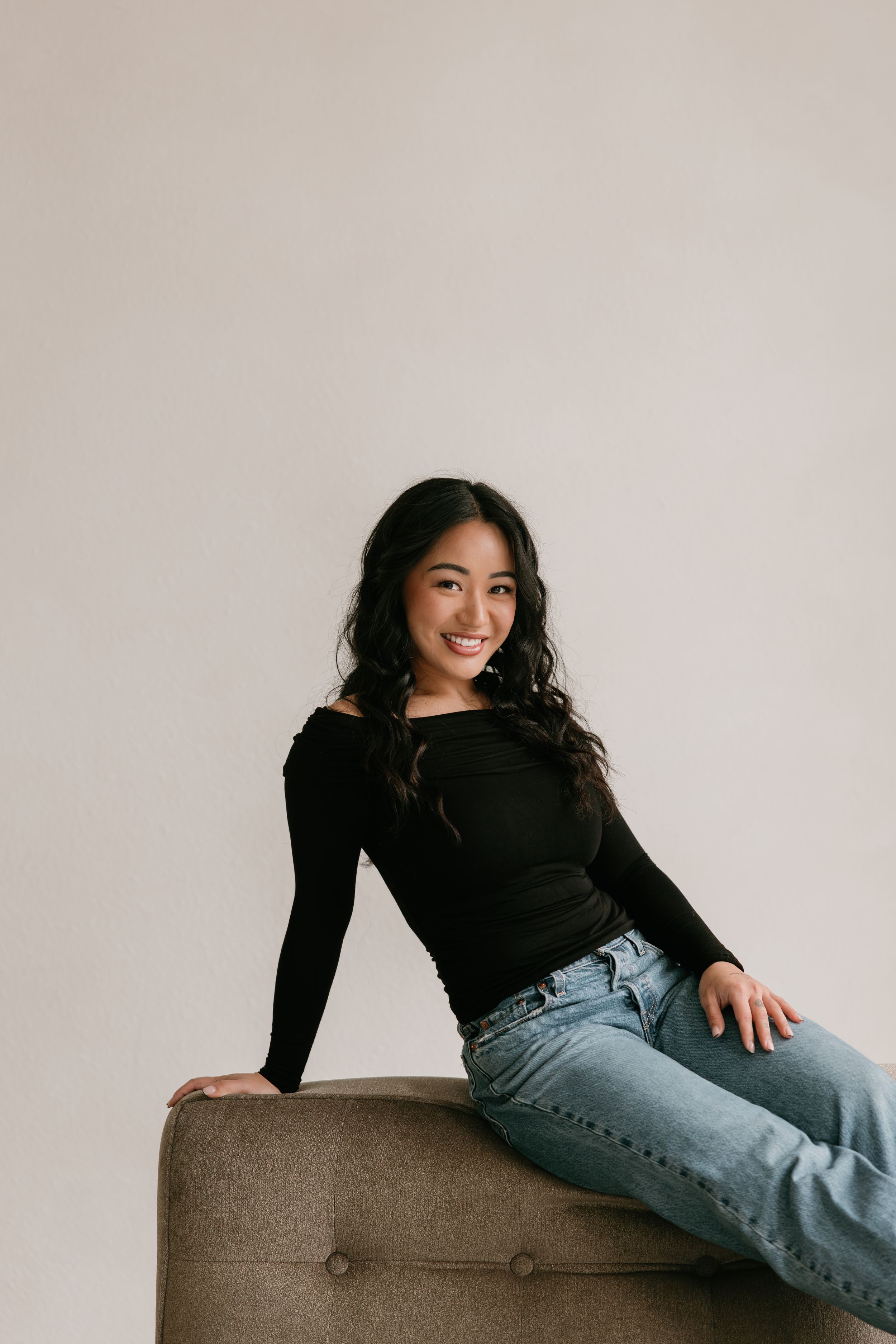 A person in a black long sleeve top and blue jeans lounges casually across a brown ottoman in a studio setting.