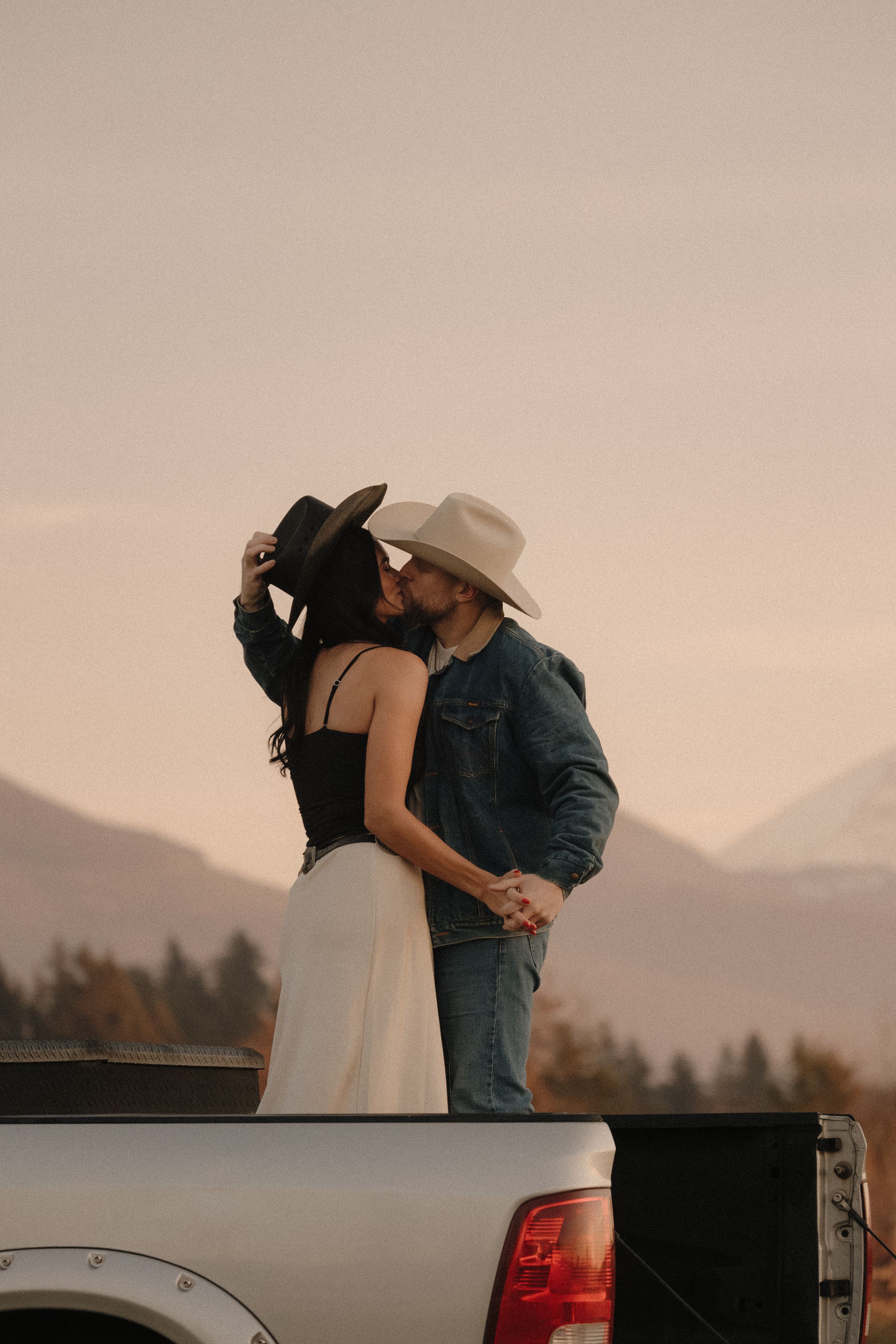 Couple in cowboy hats kissing on a truck bed at sunset with mountains in the background.