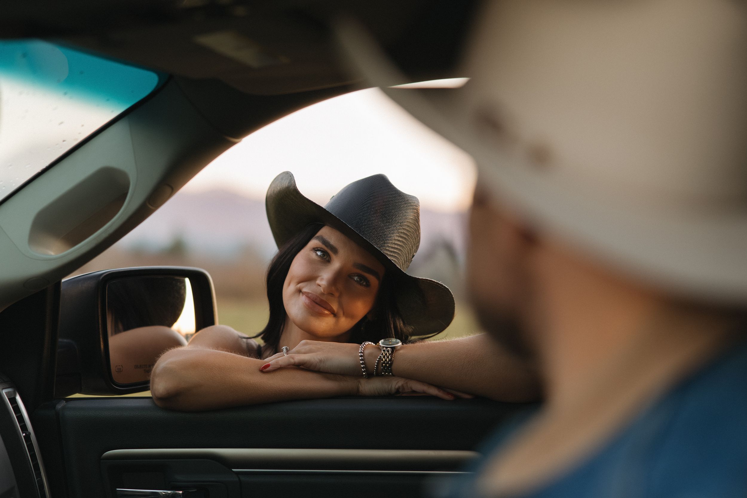 Woman in cowboy hat leaning on car window smiling at man in white hat during golden hour.