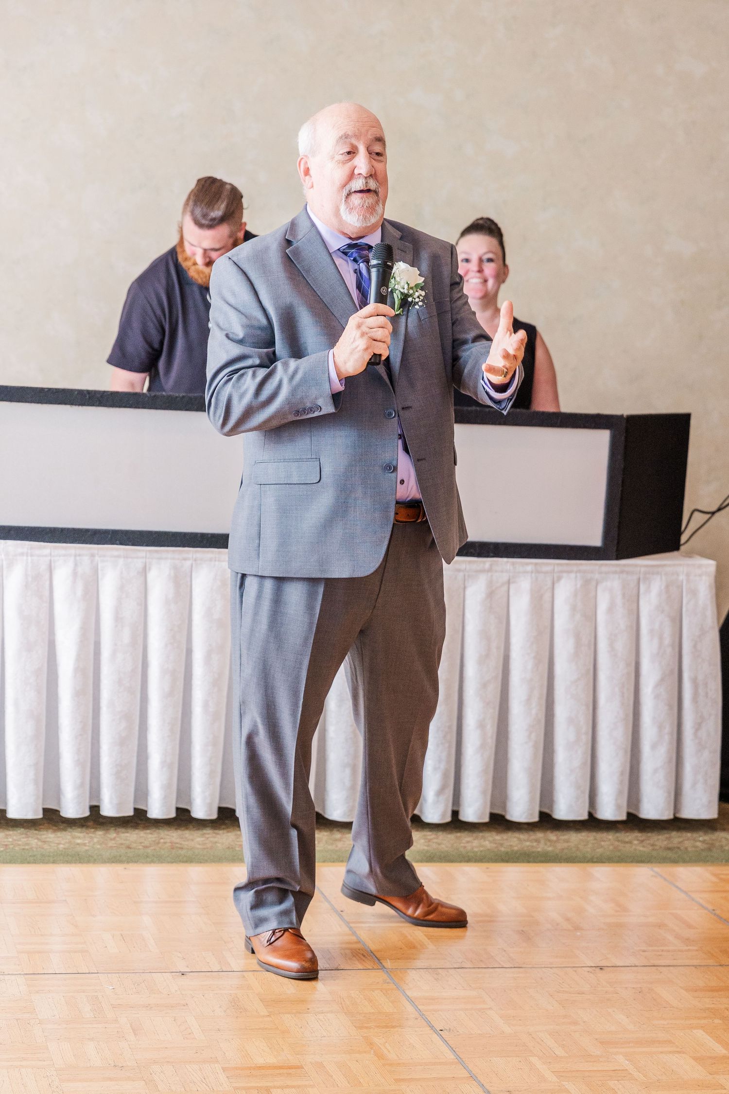 Person in grey suit speaking into microphone at formal event with others in background.