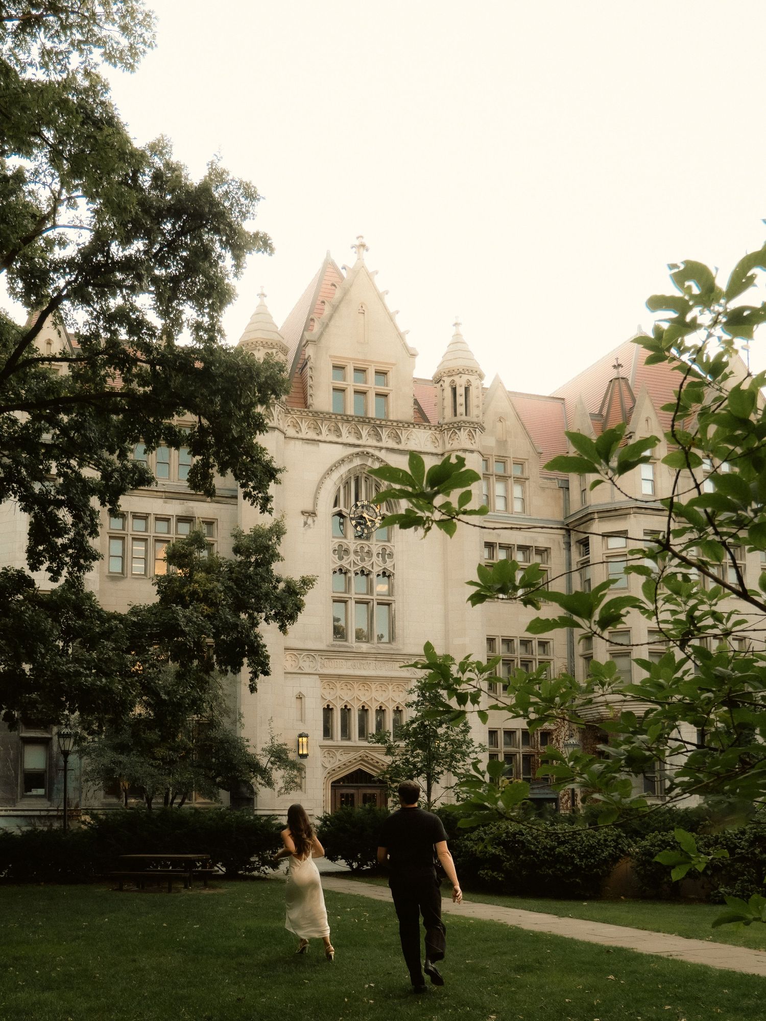 A couple walks along a path by a grand Gothic Revival university building with ornate stone architecture at dusk.