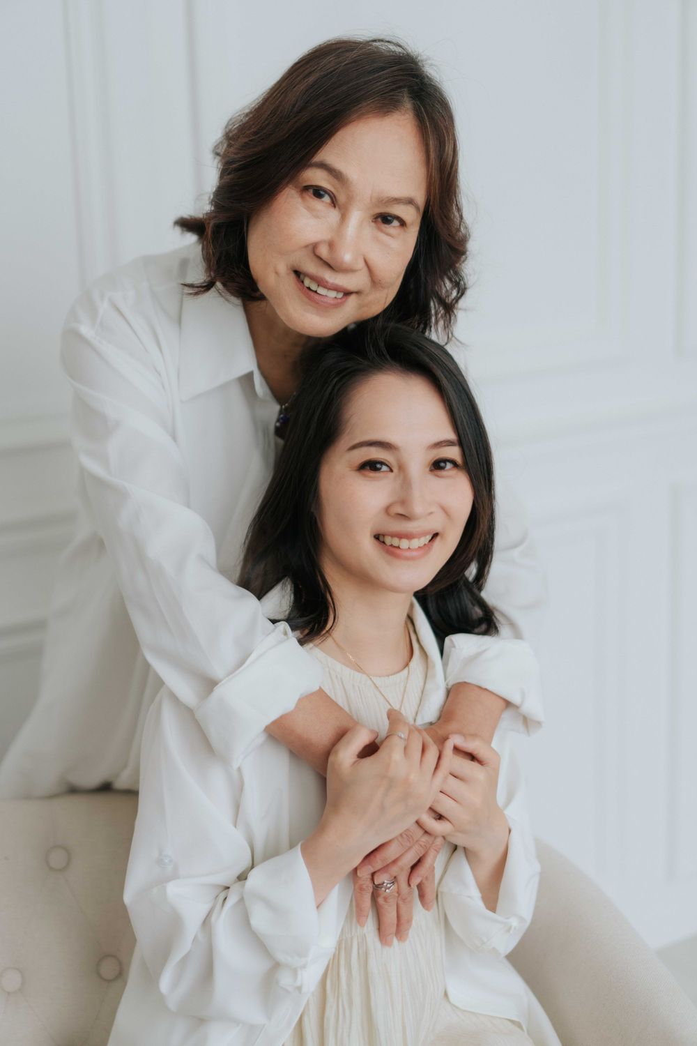 Two people in white clothing share intimate embrace while sitting on light colored furniture.