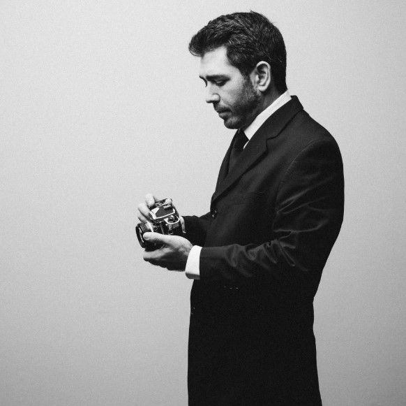 Man in dark suit stands in profile view looking down at something in his hands in black and white photo series.