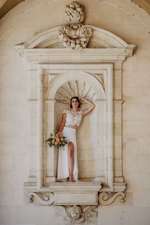 Bride in two-piece wedding gown poses in ornate stone architectural niche with bouquet.