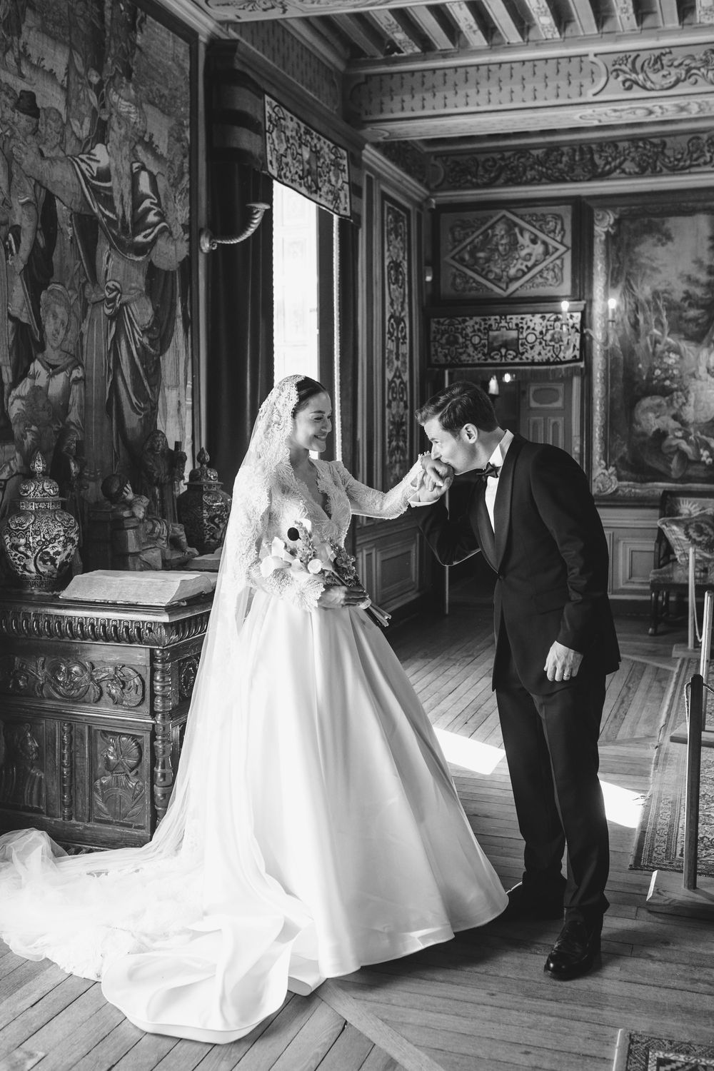 Black and white wedding photo of groom kissing bride's hand in an ornate château room with tapestries.